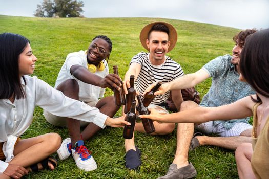 Multiracial happy and smiling friends having fun together in a park toasting with beers.