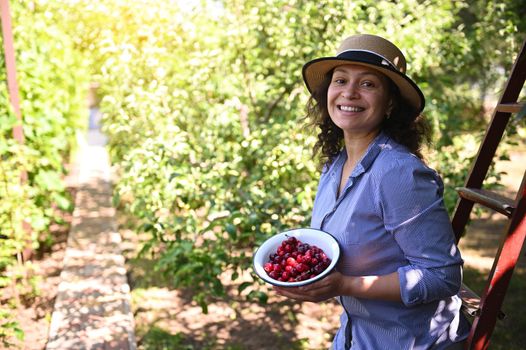 Delightful woman, eco farmer smiling toothy smile, looking at camera, holding a bowl with freshly harvested cherries