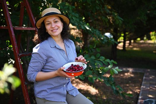 Beautiful Caucasian woman sitting on a ladder and eating sweet ripe cherries in a cherry orchard looking at camera