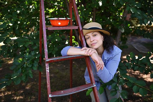 confident beautiful Caucasian woman, farmer in straw hat, leaning on a ladder in a cherry orchard and looking at camera
