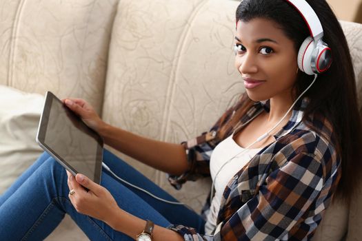 A dark-skinned girl in headphones with tablet sits on sofa