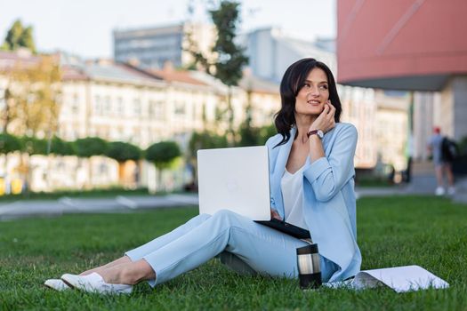 Beautiful young business woman sitting on the grass near the office center, working on laptop