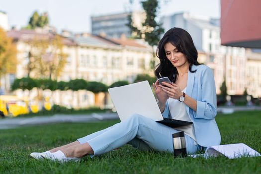 Beautiful young business woman sitting on the grass near the office center, working on laptop