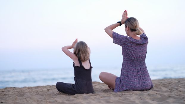 Mother and daughter do yoga meditate in lotus position on beach.