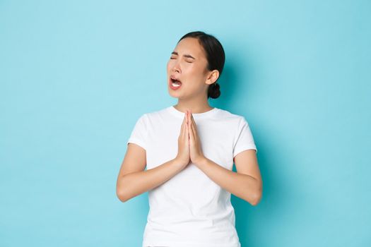Distressed uneasy asian girl pleading and crying, begging for help with exhausted upset expression, need favour, got in trouble and supplicating, feeling desperate over blue background