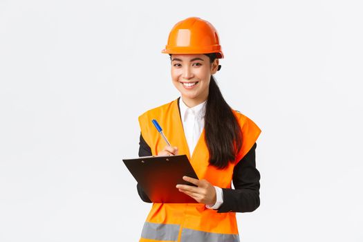 Smiling satisfied asian female construction engineer leading inspection at enterprise, wearing safety helmet and reflective jacket, writing down notes and looking pleased result, white background