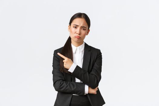 Disappointed gloomy asian female entrepreneur losing, failing job, standing in suit, pouting and pointing finger left at failure. Upset businesswoman sharing bad news, white background