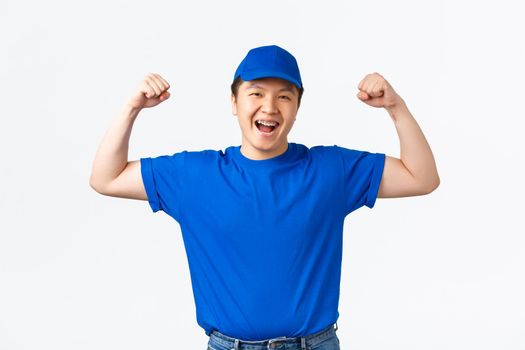 Cheerful smiling asian delivery man flex biceps and guarantee safety of packages. Courier in blue uniform show-off his strong hands, carry parcels to clients homes, standing white background