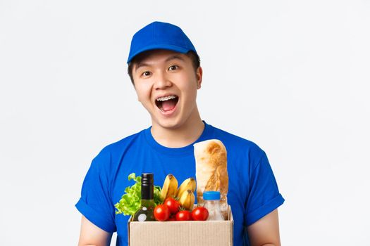 Online shopping, food delivery and shipment concept. Close-up of excited smiling young asian courier in blue uniform cap and t-shirt, handing groceries order to customer, white background