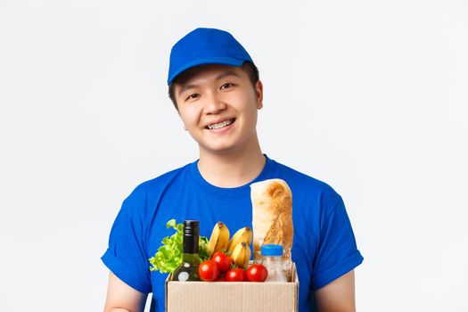 Online shopping, food delivery and shipment concept. Close-up of smiling pleasant asian male courier, wearing blue uniform, handing box with grocery order to client, standing white background