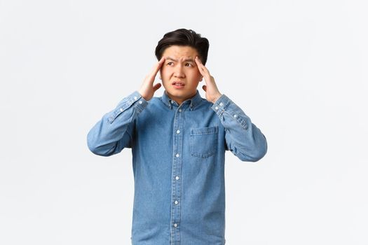 Troubled and uneasy asian man looking perplexed, having complicated situation. Guy with headache touching head and looking away, suffering painful migraine, standing white background