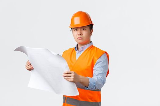 Building sector and industrial workers concept. Serious-looking confident asian construction manager, engineer in helmet and reflective jacket looking at blueprint, checking layout, white background