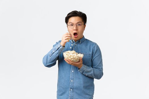 Leisure, lifestyle and people concept. Thrilled and amazed asian guy watching awesome movie or TV series, eating popcorn and looking astounished, standing white background