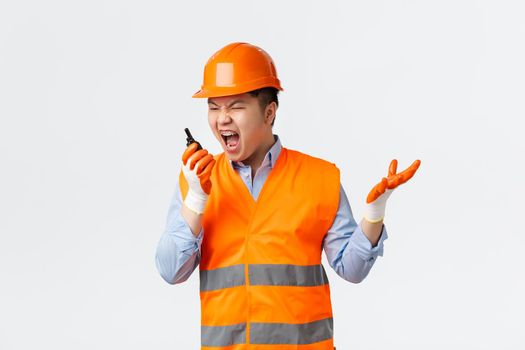 Building sector and industrial workers concept. Angry and pissed-off asian chief engineer scolding workers for mistakes, shouting in walkie-talkie furious, standing in helmet and reflective clothing