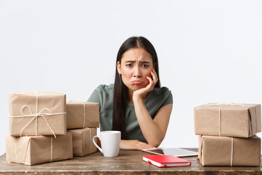 Small business owners, startup and e-commerce concept. Sad and gloomy asian businesswoman pouting as not have clients, waiting for orders, looking distressed sitting near piles of boxes