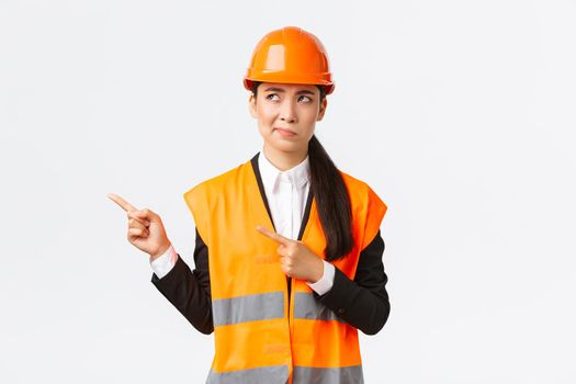 Building, construction and industrial concept. Indecisive asian female architect being unsure, wearing reflective clothing and helmer, smirk as pointing and looking upper left corner puzzled