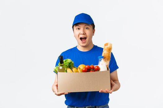 Online shopping, food delivery and shipment concept. Amazed smiling young asian male courier, wearing uniform and handing box with fresh groceries to customer, bring order, white background