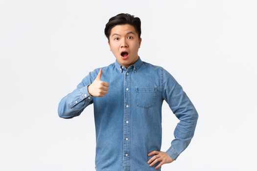 Surprised and impressed asian male student open mouth fascinated, showing thumbs-up astounded, saying wow, rate great work, saying well done or good job, standing white background