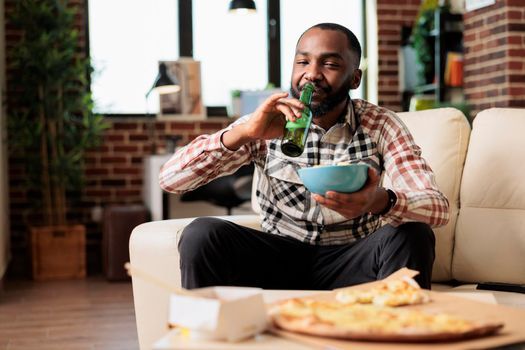 Cheerful adult drinking alcoholic beer from bottle and holding bowl of chips