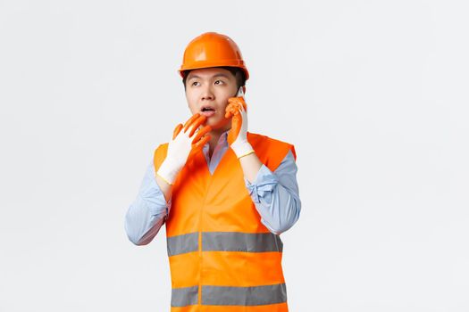 Building sector and industrial workers concept. Thoughtful asian engineer working construction area, wearing helmet and reflective clothes, answering question over mobile phone, thinking