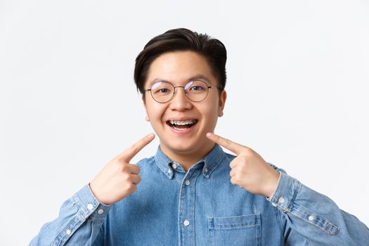 Orthodontics and stomatology concept. Close-up of satisfied asian guy, dental clinic client smiling happy and pointing at his dental braces, standing white background, recommend quality
