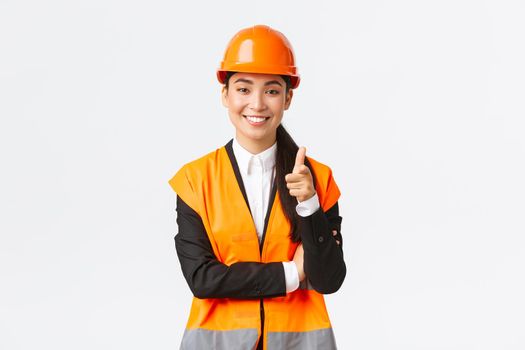Successful and confident smiling asian female architect pointing finger at camera, wearing safety helmet and reflective jacket, introduce construction project, industrial woman inviting clients