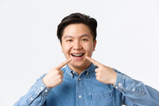 Orthodontics and stomatology concept. Close-up of happy smiling asian man pointing fingers at dental braces on teeth with pleased expression, recommend dentist clinic, standing white background