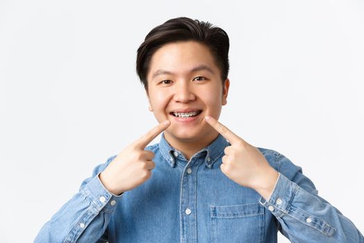 Orthodontics and stomatology concept. Close-up shot of happy smiling asian man recommend dental clinic, pointing fingers at teeth braces with satisfied expression, white background