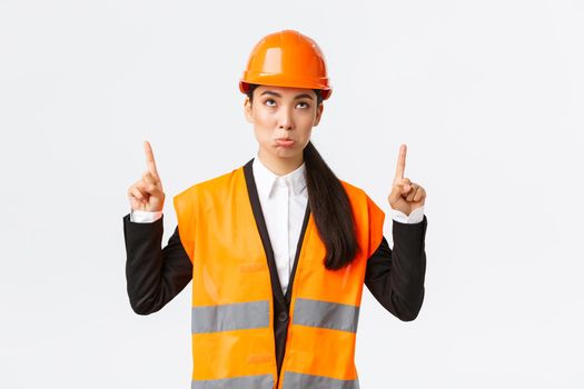 Building, construction and industrial concept. Disappointed gloomy asian female engineer, architect in safety helmet and reflective clothing pouting displeased as looking and pointing fingers up