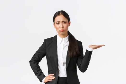 Disappointed and gloomy female sales manager, real estate agent in black suit having bad day, pouting and look upset while showing something, holding hand to the right, white background