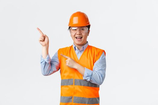 Building sector and industrial workers concept. Smiling cheerful asian architect, chief engineer in safety helmet and reflective jacket, pointing fingers upper left corner, showing banner
