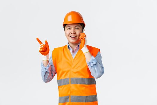 Building sector and industrial workers concept. Smiling asian engineer, construction manager in reflective clothings and helmet pointing finger left while talking on mobile phone, white background