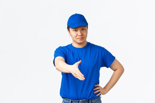 Friendly cheerful asian courier in blue uniform smiling confident, extend hand for handshake, greeting customer, making deal. Delivery man saying hello and standing white background