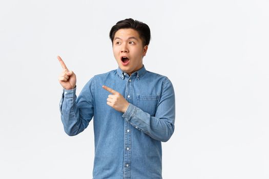 Impressed and astounded young asian man react to fantastic news, open mouth fascinated, pointing and looking upper left corner amazing advertisement banner, standing white background