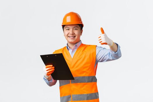 Building sector and industrial workers concept. Smiling satisfied asian chief engineer, architect making inspection showing thumbs-up in approval, holding clipboard with documents, wear helmet