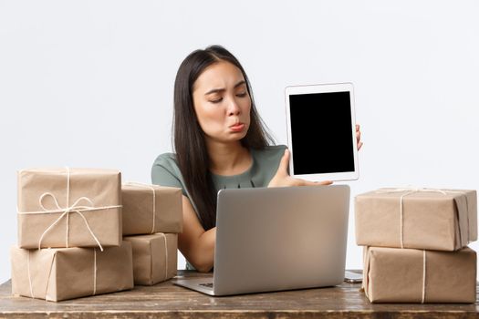Small business owners, startup and e-commerce concept. Disappointed asian woman pouting upset, showing bad stats on digital tablet screen, using laptop, packing orders in boxes for shipping