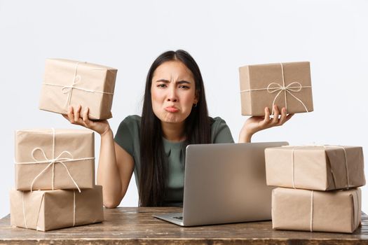 Small business owners, startup and e-commerce concept. Pouting tired asian woman managing online store, loaded with work, sitting with laptop and piles of boxes waiting for shipping, grimacing upset