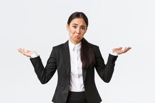 Clueless upset young asian female office worker, manager in black suit, shrugging with hands spread sideways and pouting unaware, dont know, cant understand, standing useless and indecisive