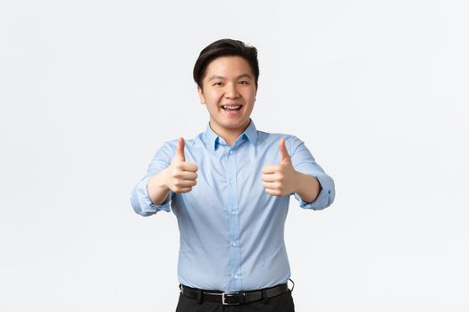 Business, finance and people concept. Enthusiastic handsome asian male office worker, employee with teeth braces, showing thumbs-up in approval, recommend company, guarantee quality