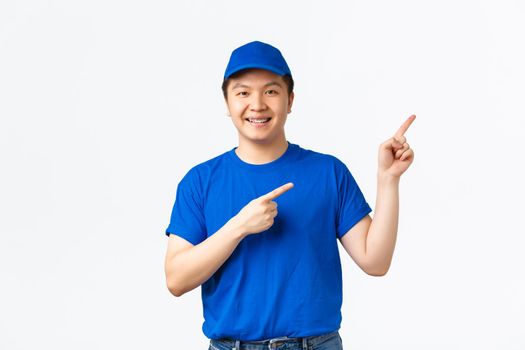 Friendly-looking cheerful asian courier with braces, wearing blue uniform pointing fingers upper right corner and smiling, inform clients about shipment rules or special discounts, white background