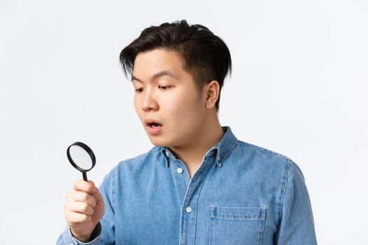 Close-up of young asian guy looking curious and focused, searching for something, looking through magnifying glass down, open mouth intrigued, standing white background