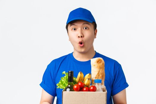 Online shopping, food delivery and shipment concept. Amazed and wondered asian male courier in blue uniform looking astonished, saying wow, carry box with groceries to customer house