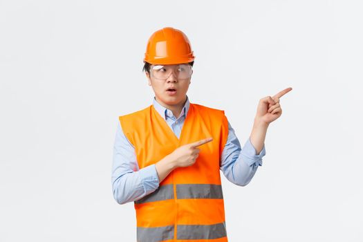 Building sector and industrial workers concept. Concerned and doubtful asian male construction manager, engineer in helmet and reflective clothing pointing upper right corner hesitant