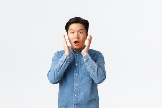 Surprised and impressed asian guy react to big announcement, holding hands near face and gasping, stare at something unbelievable, standing astounded over white background