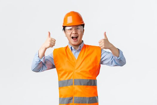 Building sector and industrial workers concept. Smiling asian male engineer, architect or builder in safety helmet and reflective clothing showing thumbs-up, assure everything alright