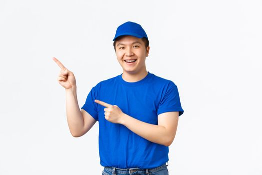 Cheerful young asian male courier in blue uniform pointing fingers upper left corner, advertisement about shipping company, parcels shipment. Cute delivery guy promote something