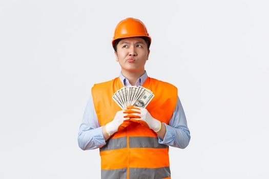 Building sector and industrial workers concept. Thoughtful and perplexed asian construction manager in helmet and reflective clothing pondering while holding money, white background