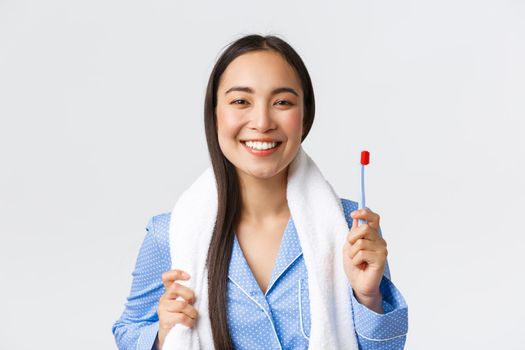 Daily routine, morning and hygiene concept. Upbeat smiling beautiful asian girl in blue pajama getting ready to shower, holding towel and toothbrush, grinning white teeth, white background