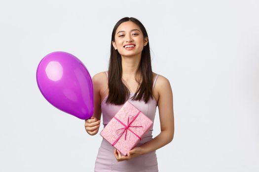 Celebration, party and holidays concept. Beautiful asian girl in dress, celebrating birthday, holding balloon and b-day gift, smiling and thanking for surprise, white background