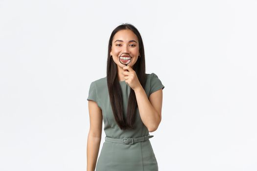 Healthcare, beauty and stomatology concept. Funny asian female with perfect white smile holding magnifying glass over teeth and grinning upbeat, standing white background